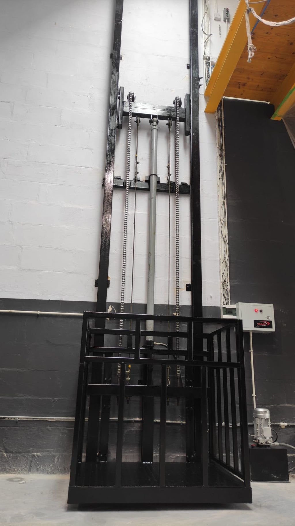 Front view of a black guide rail cargo lift showing the platform, lifting chains and hydraulic cylinder.
