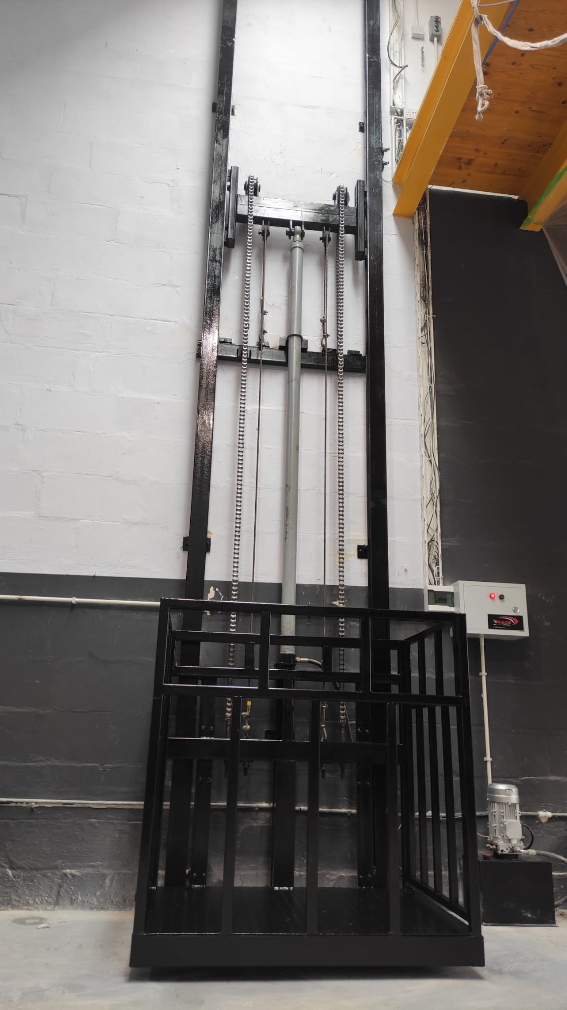 Front view of a black guide rail cargo lift showing the platform, lifting chains and hydraulic cylinder.