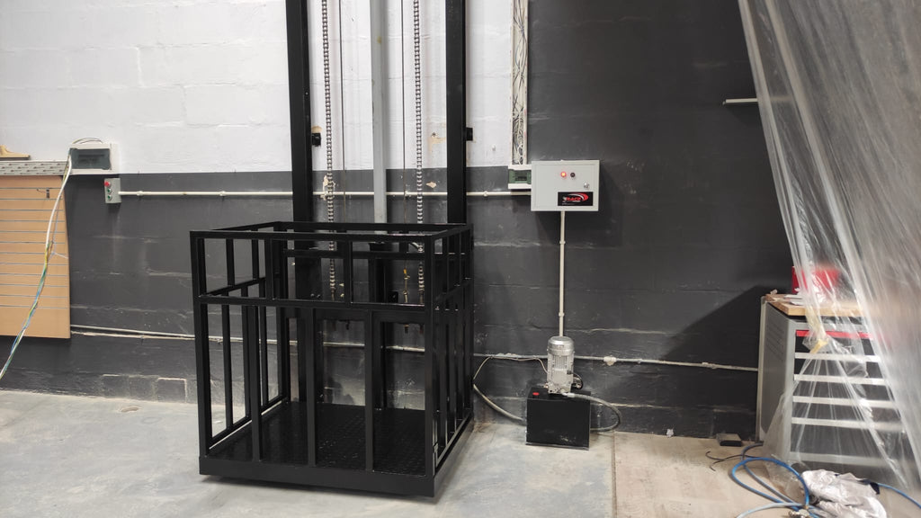 Black guide rail cargo lift at floor level with platform guardrails, a wall-mounted control box and pump unit