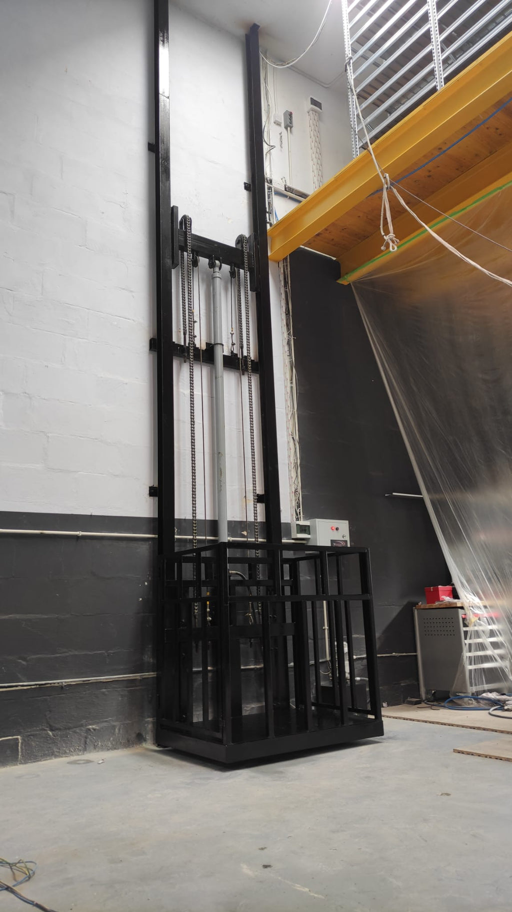 Angled view of a black guide rail cargo lift installed beside a mezzanine, with the platform at floor level.